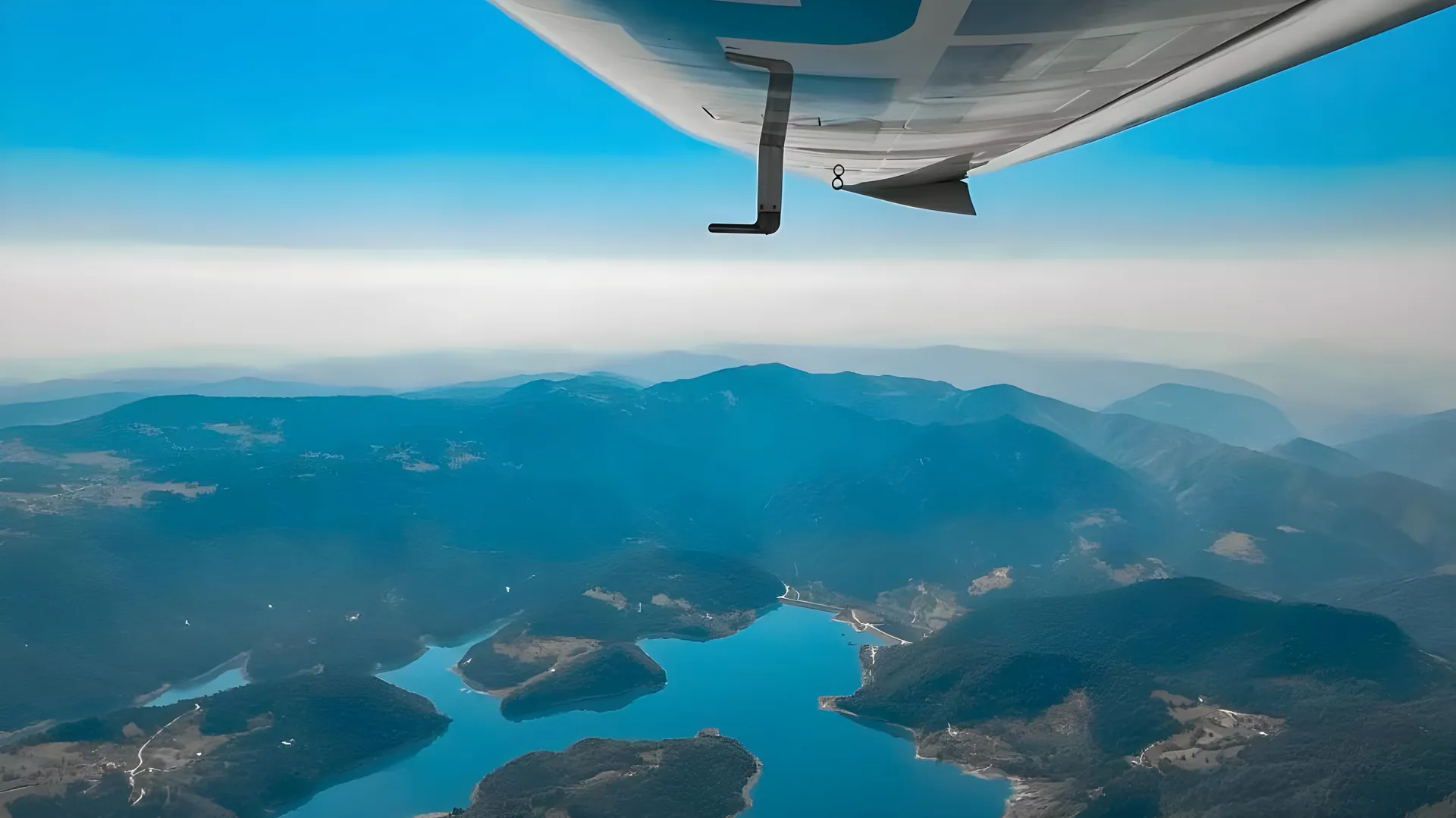 View from under the wing of a small training aircraft, flying over forested mountains and a winding blue lake
